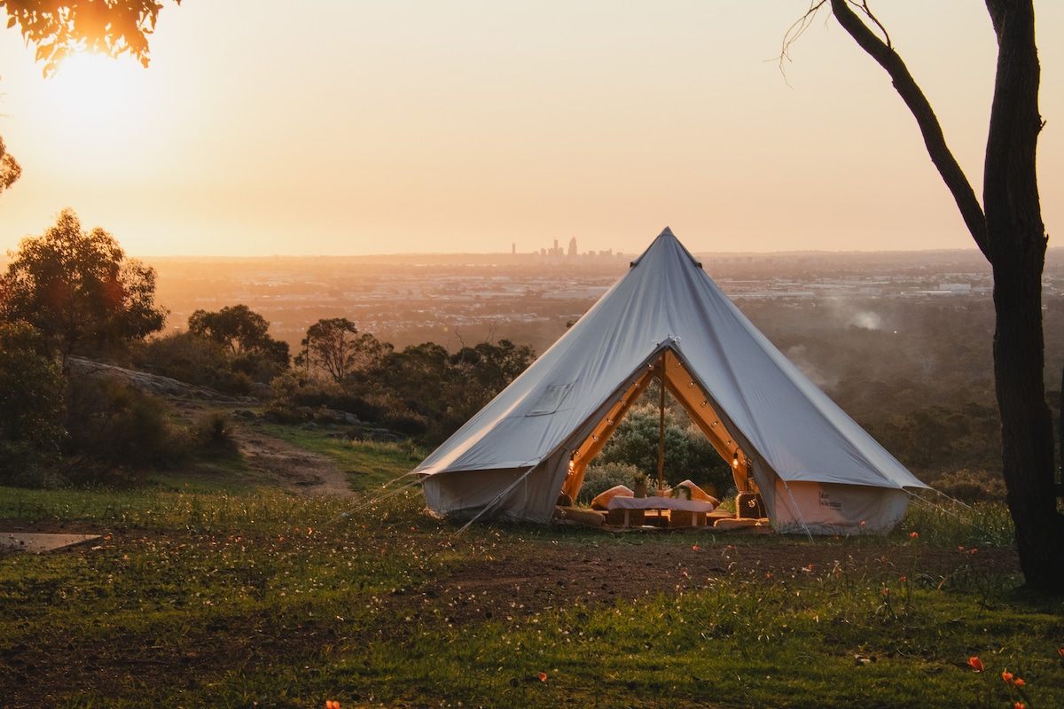 A large, tent-like structure is situated on a grassy hillside, offering a view of the distant city skyline against a sunset backdrop. The tent's entrance is open, revealing a cozy setup inside, while surrounding trees frame the serene landscape.