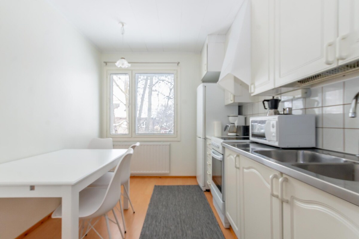 A clean and well-lit kitchen space features white cabinetry and stainless steel appliances. A dining table is accompanied by four white chairs, allowing for meal preparation and dining. A window invites natural light, with views of trees outside creating a refreshing ambiance.