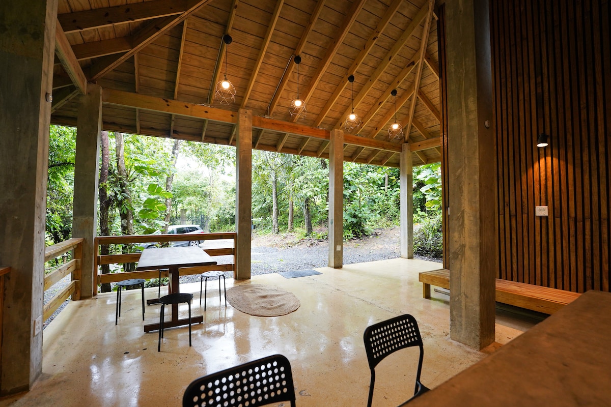 An open-air space is featured, supported by concrete pillars and a wooden roof. Large windows allow light to fill the area, which includes a dining table with chairs. Lush greenery is visible outside, highlighting the natural surroundings.