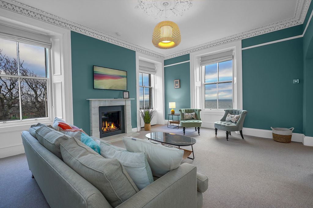 An elegantly furnished sitting room features a marble fireplace with a living flame gas fire, complemented by a wall-mounted Smart TV. Large windows provide ample natural light and sea views. Comfortable seating arrangements include a large sofa and accent chairs, with a coffee table at the center.