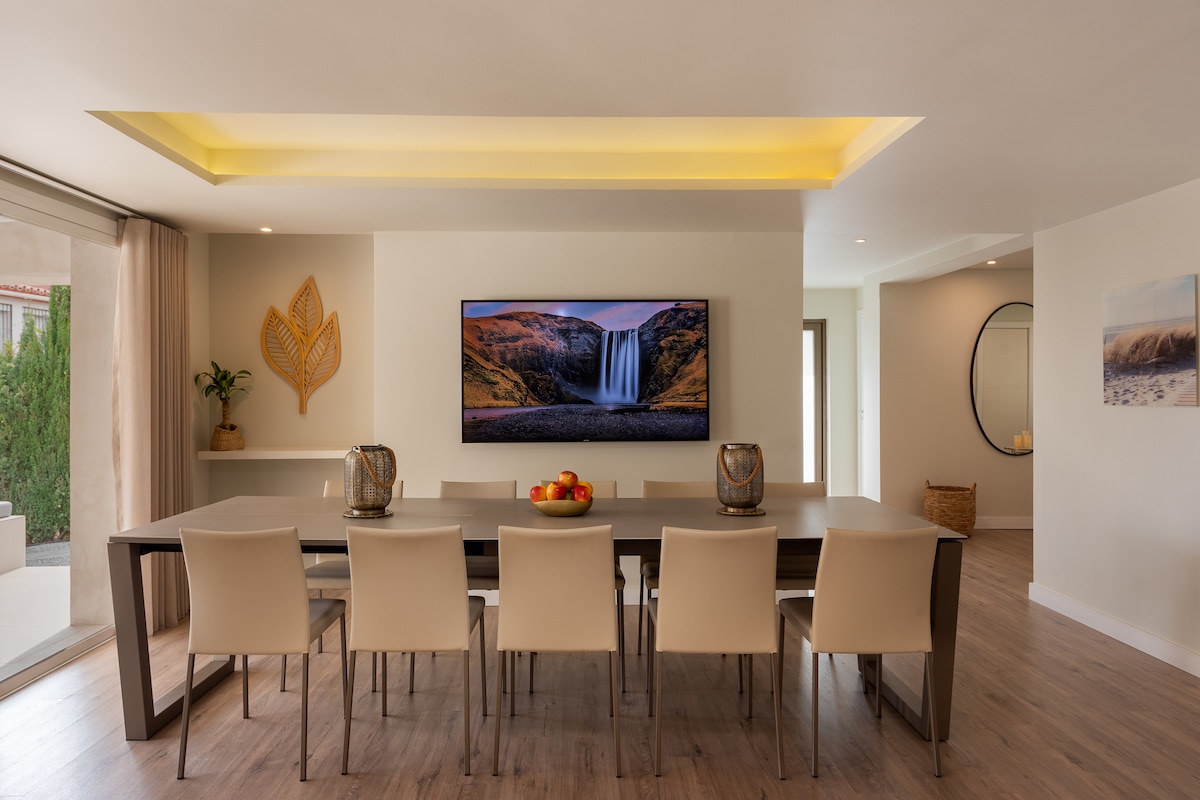 A spacious dining area features a long table with seating for twelve, illuminated by soft overhead lighting. A large flat-screen TV is mounted on the wall, flanked by decorative elements, while natural light enters through expansive sliding glass doors leading to the terrace.