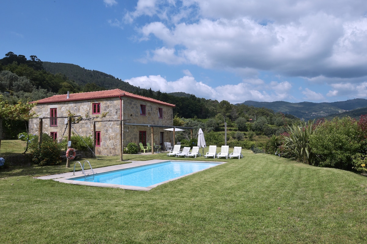 A tranquil exterior view of a stone house surrounded by greenery and mountains. A clear swimming pool is framed by a spacious lawn, dotted with sun loungers. Bright red accents are visible on the windows and doors, enhancing the rustic charm of the property.