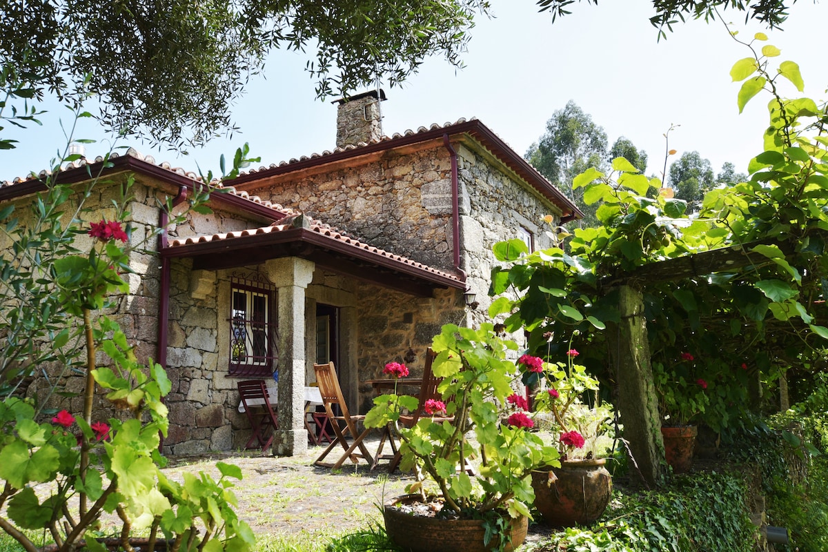 A rustic stone house is surrounded by lush greenery and vibrant flowers. The structure features a tiled roof and wooden accents. Comfortable seating is positioned outside, inviting relaxation in the garden space adorned with potted plants. Sunlight filters through the leaves, creating a serene atmosphere.