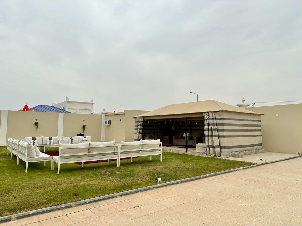 An outdoor majlis features a spacious seating area, arranged around a central rug, with white couches and colorful cushions. A large tent provides shelter, and the surrounding green lawn enhances the relaxed atmosphere. Soft lighting fixtures are visible along the perimeter.