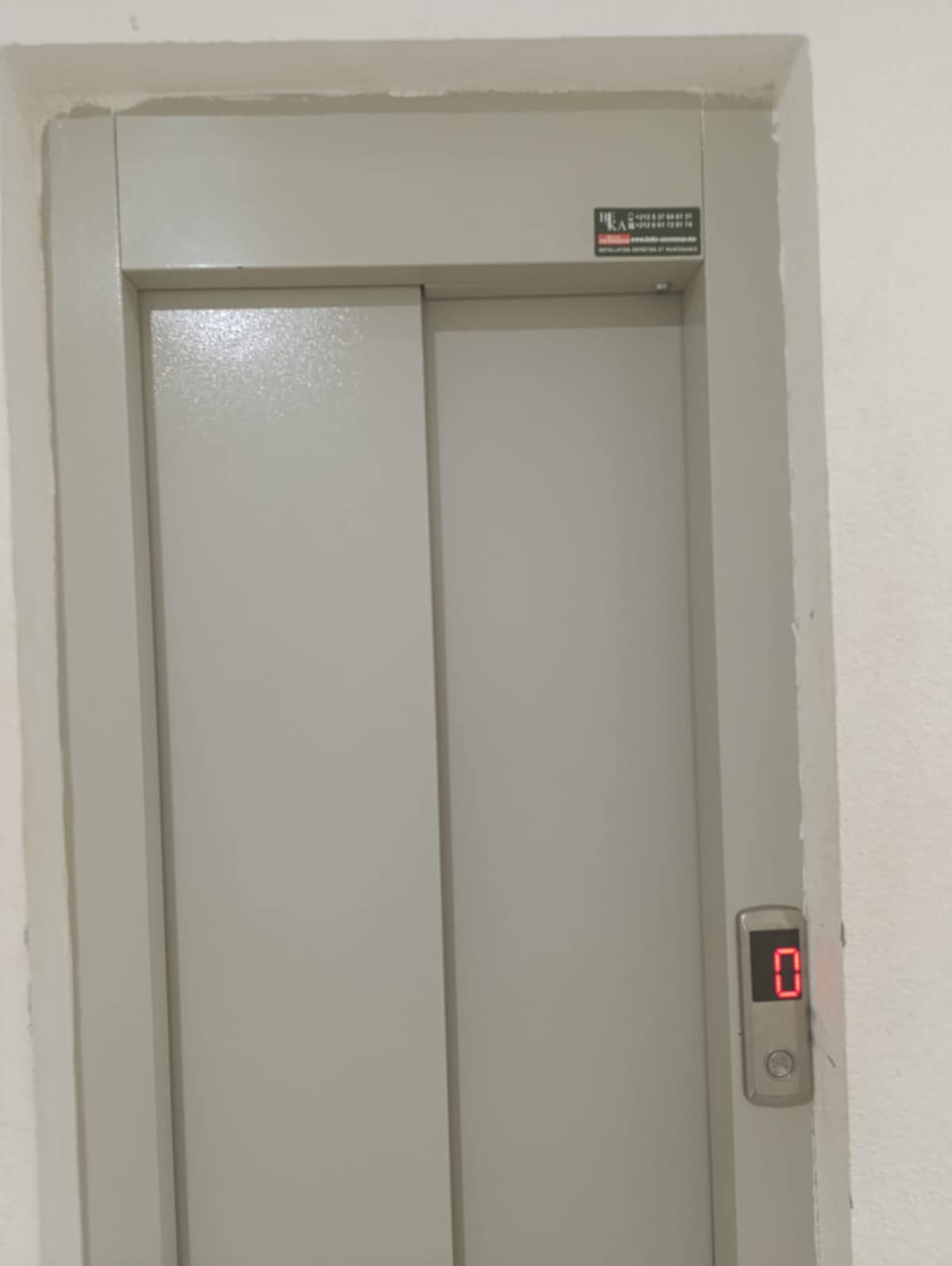 A neutral-colored elevator door is shown, featuring a simple design with smooth surfaces. A digital display indicates the current floor number, while a button panel is positioned beside the door for easy access.