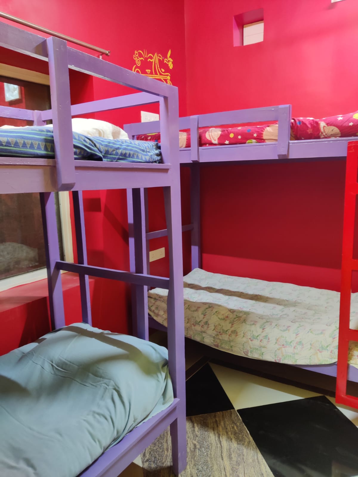 A vibrant room featuring two sets of bunk beds with colorful frames in purple and red. Each bed is equipped with bedding, while the walls are painted in a lively red shade. A window allows natural light to fill the space.
