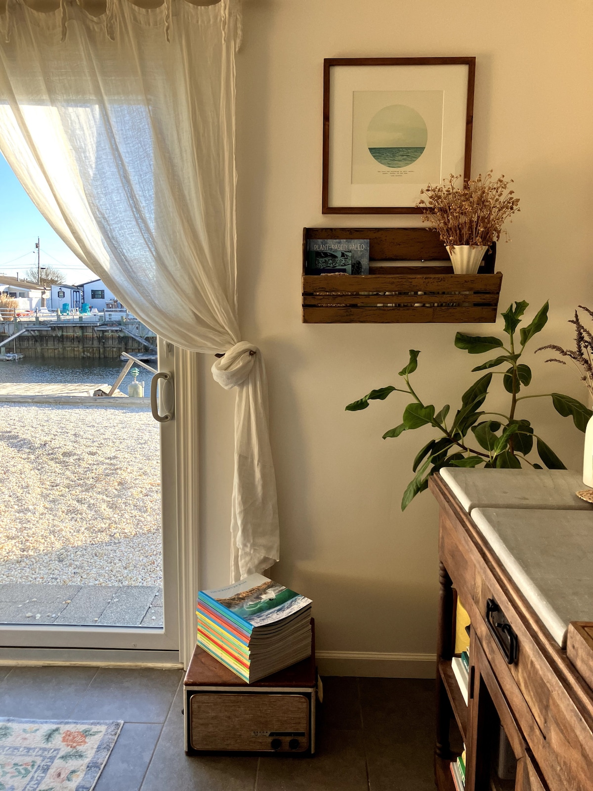 Natural light floods the entry space, highlighting a neatly arranged wooden crate with succulents and photographs. The soft, flowing curtain gently frames the view of the outdoor area, where a glimpse of the water adds a sense of tranquility. A stack of colorful books is positioned below.