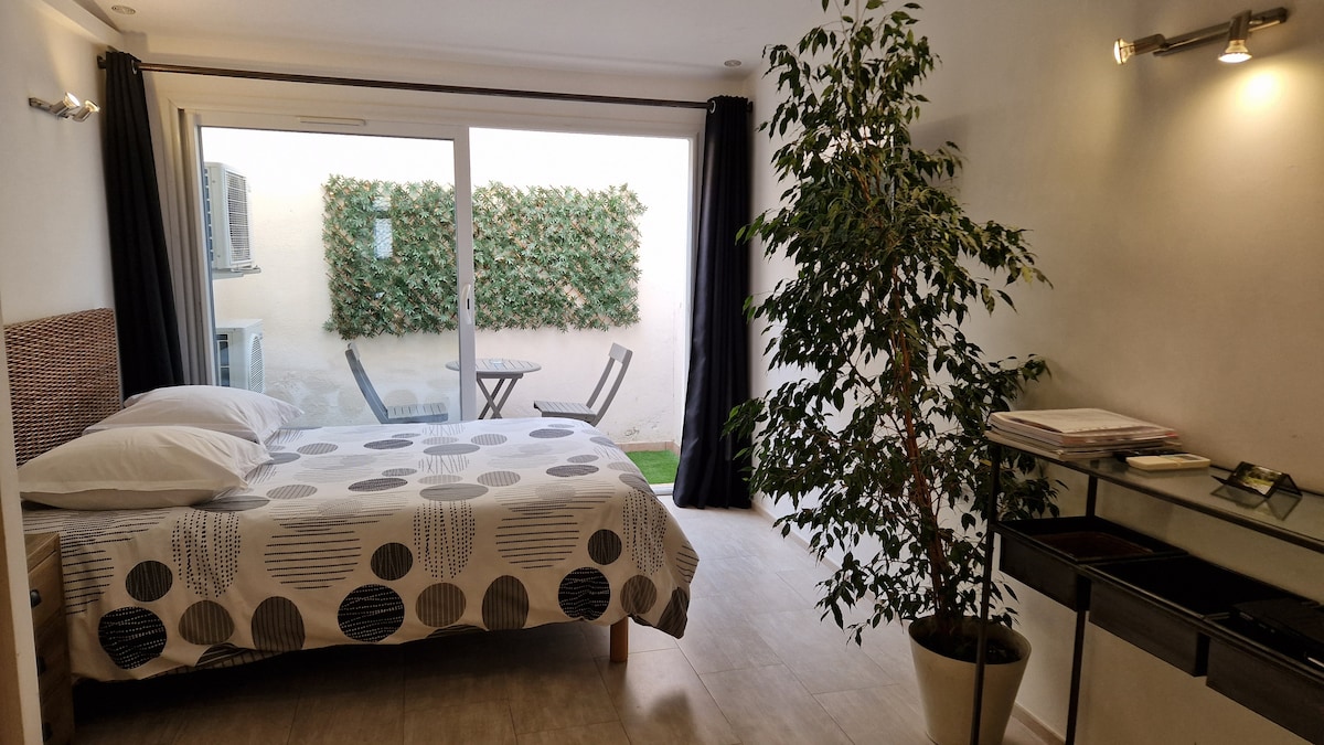 A bright and airy studio is furnished with a spacious bed adorned with patterned linens. A small terrace with two chairs and a table is visible through glass doors, surrounded by greenery. A potted plant adds a touch of nature to the interior.