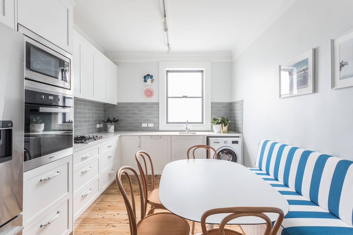 A modern kitchen is equipped with streamlined cabinetry and stainless steel appliances. A round table is set with curved chairs, complemented by a blue and white striped banquette. Natural light floods the space through a large window, highlighting the wooden flooring.