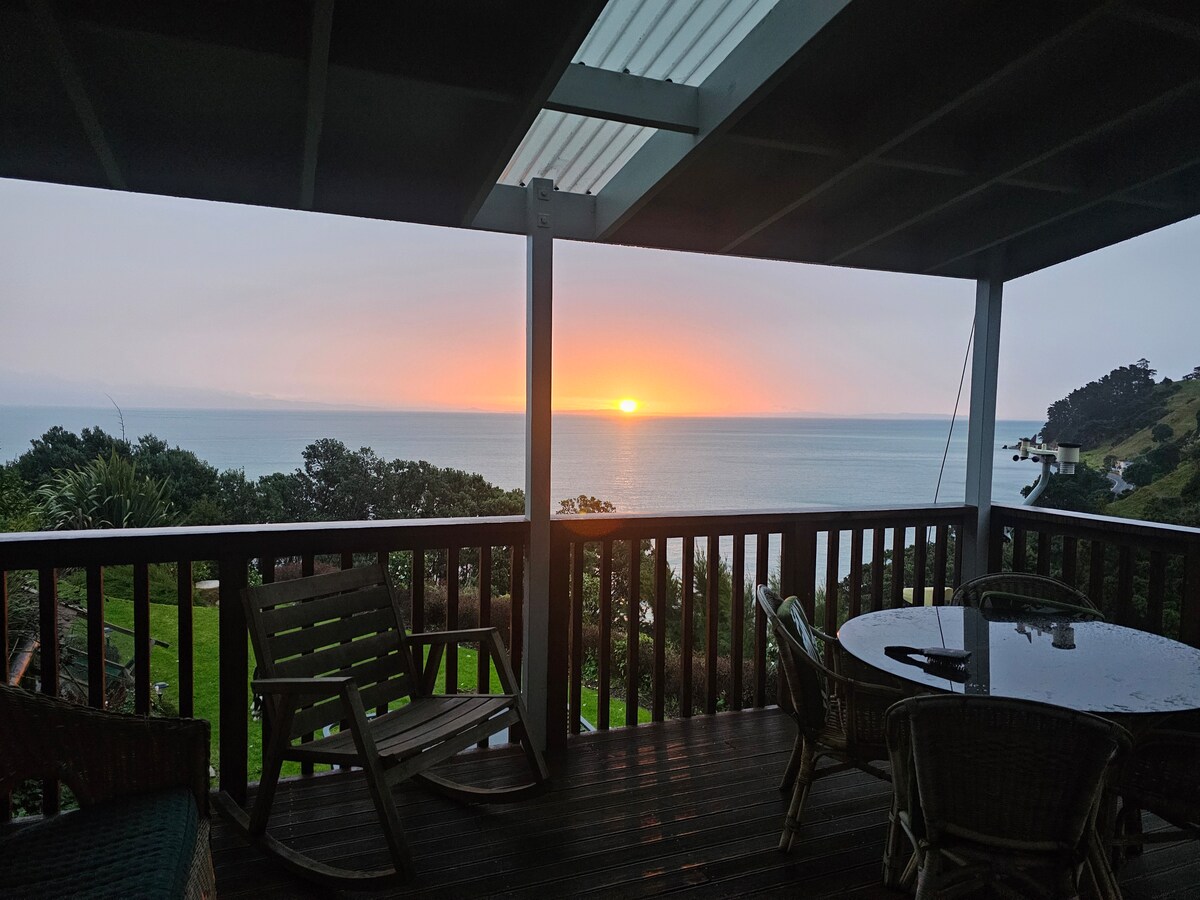 An outdoor deck is showcased, featuring a circular table and several wooden chairs. The scenery includes a breathtaking sunset over the ocean, with hues of orange and pink illuminating the horizon. Lush greenery is visible in the foreground, enhancing the coastal view.