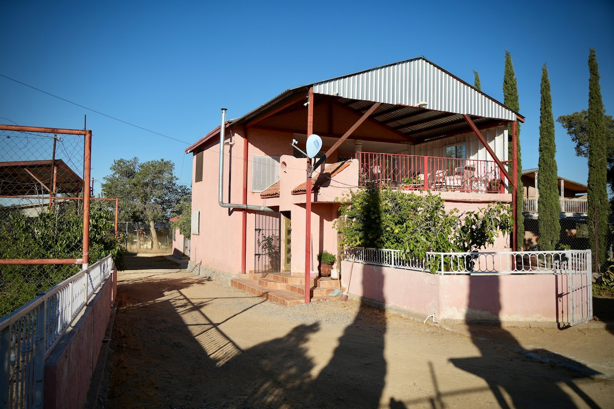 A two-story pink house is set within a spacious outdoor area, featuring a gravel driveway leading up to the entrance. Surrounding greenery provides a natural backdrop, while a balcony adorned with a railing overlooks the entrance and garden.