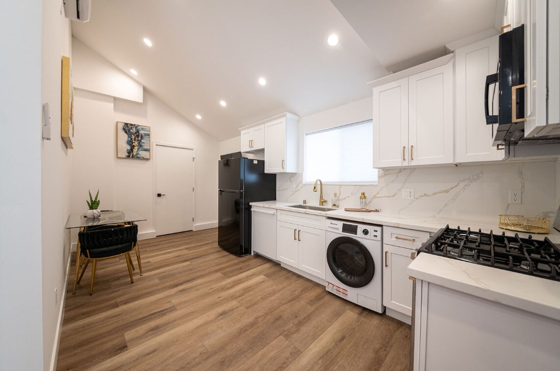 A modern kitchen is displayed, featuring white cabinetry and stainless-steel appliances. A large window provides natural light, while a small dining table with black chairs is positioned to the side. Countertops exhibit a marble-like finish, and a washing machine is integrated into the space.