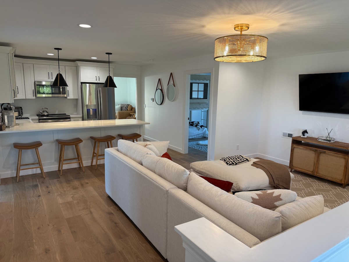 An open living area is visible, featuring a light-colored sofa adorned with decorative throw pillows. A modern kitchen is in the background, with white cabinets, bar seating for three, and pendant lighting. A television is mounted on the wall opposite, adding to the casual atmosphere.