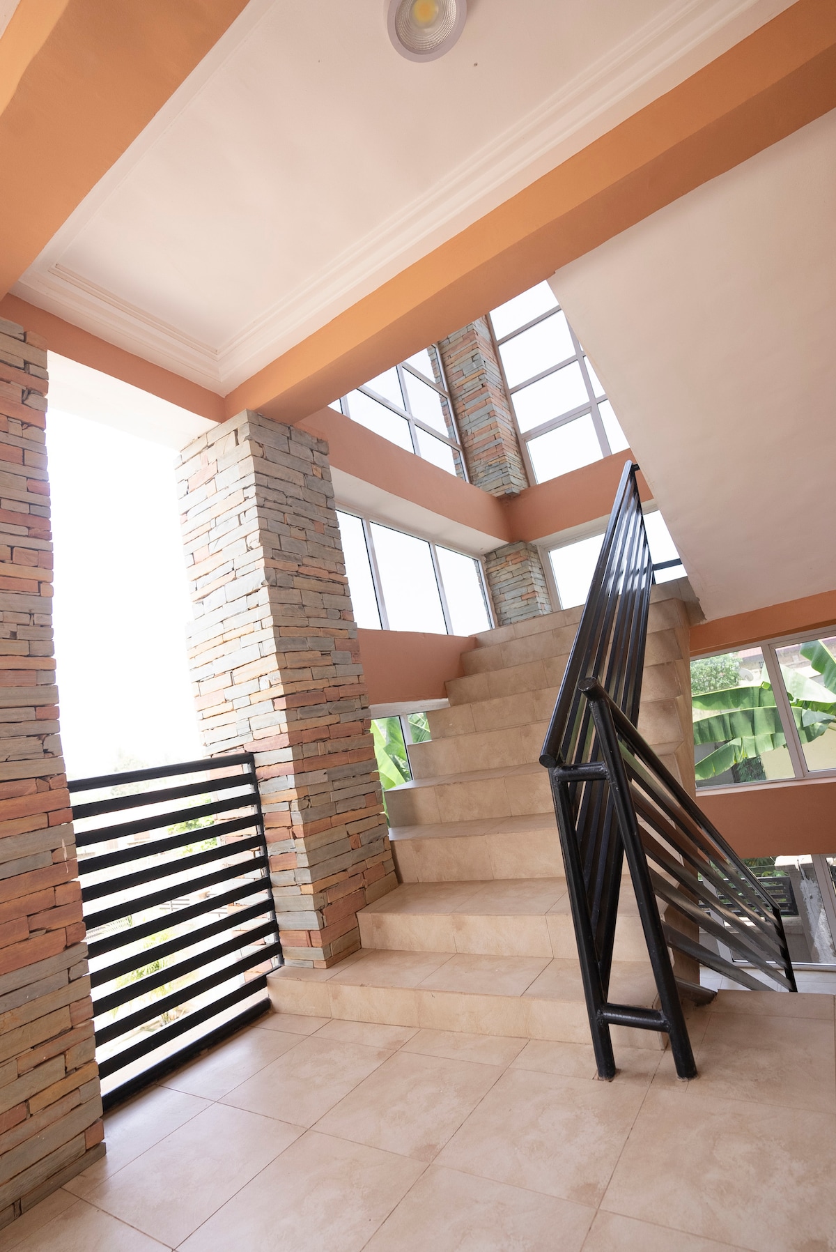 A well-lit staircase is accented by large windows, allowing natural light to enter. The walls are adorned with a mix of warm tones and stacked stone, enhancing the modern design. A sleek black railing provides safety and style along the staircase leading to upper levels.