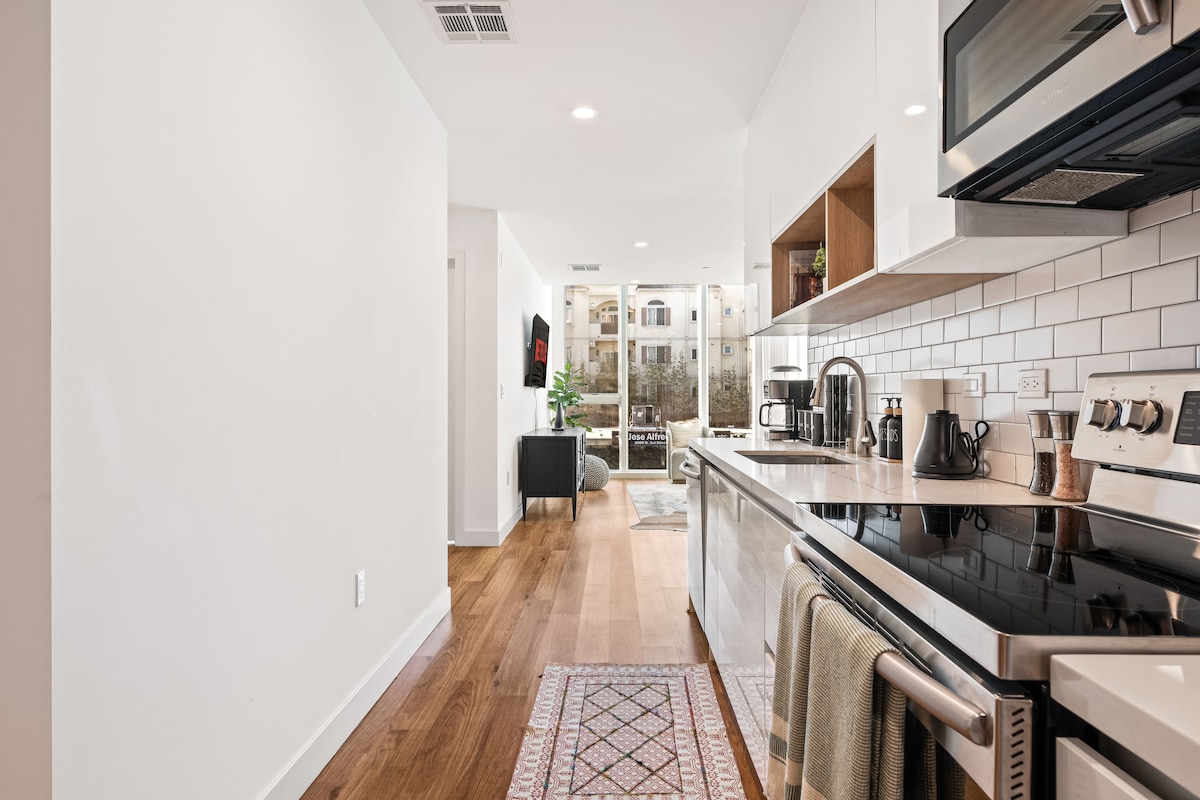 The kitchen area showcases modern appliances, including a stove and microwave, along with a sleek and clean design. White cabinetry and neutral tones complement the warm hardwood floor. A subtle area rug adds texture, leading towards a bright living space visible in the background.