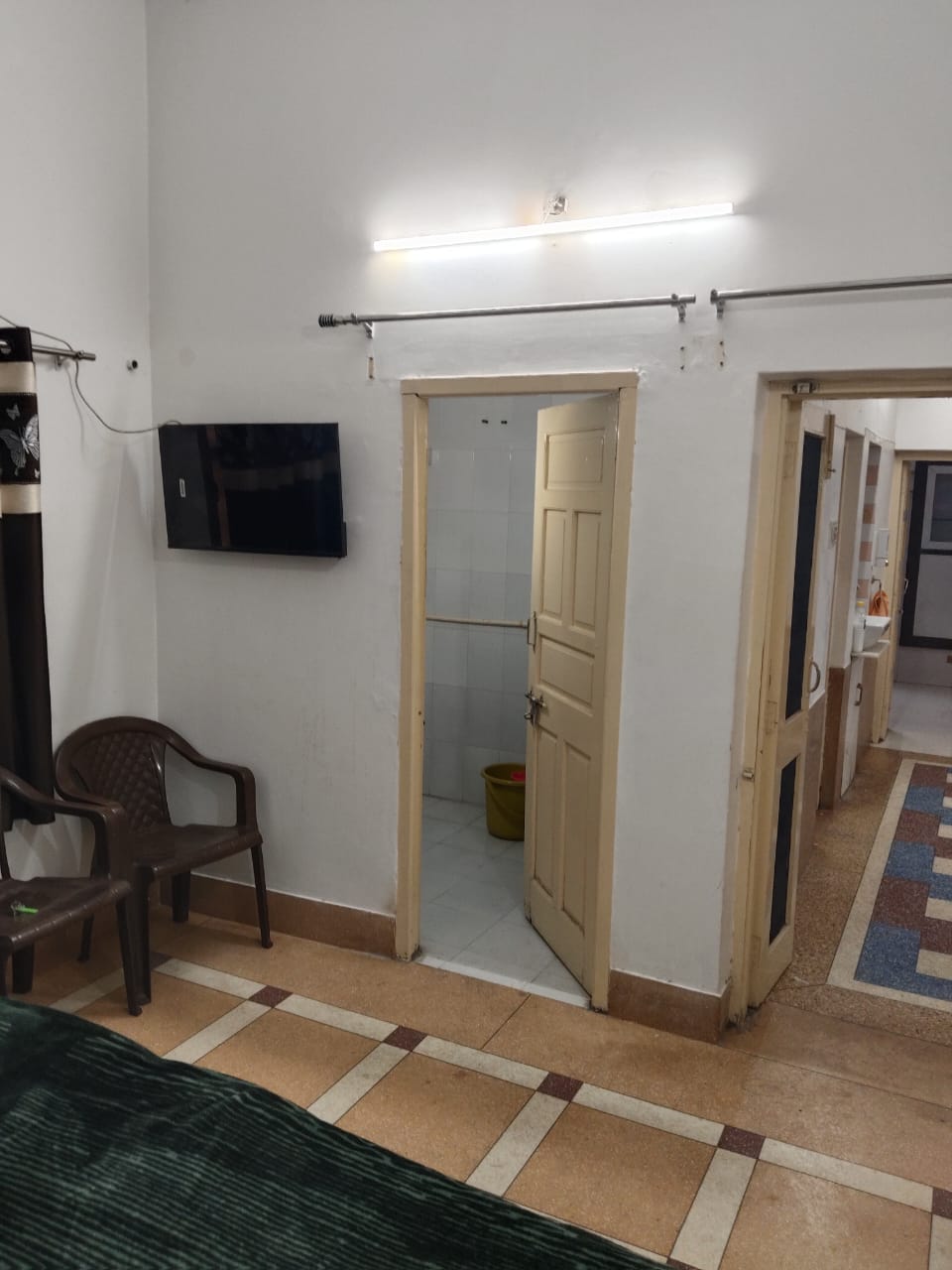 A spacious room is shown, featuring a wall-mounted television and a door leading to the attached bathroom. Two plastic chairs are positioned near the wall, while light is provided by fluorescent fixtures above. The flooring consists of beige tiles, contrasting with the dark green bed linens.