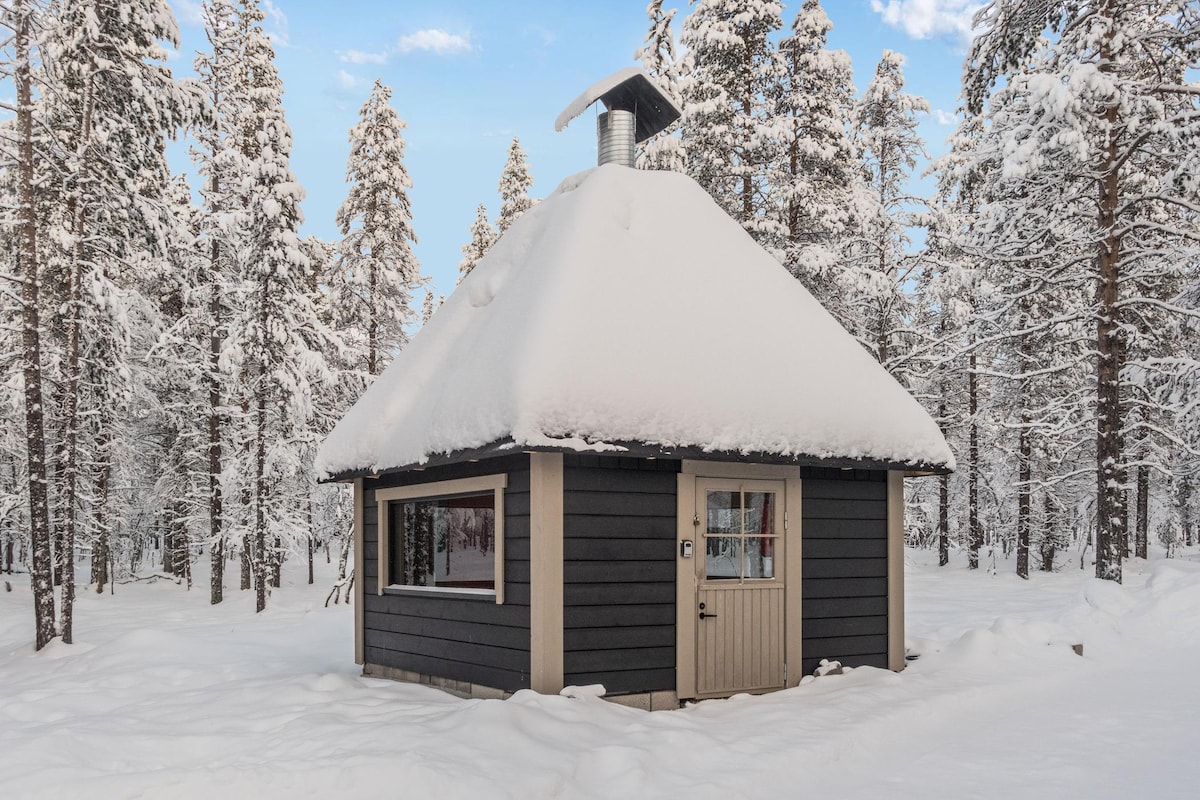A charming log cabin is surrounded by snow-covered trees, creating a serene winter landscape. The structure features a unique conical roof with a chimney, and a welcoming entrance door is visible. Soft snow blankets the ground, enhancing the peaceful atmosphere.