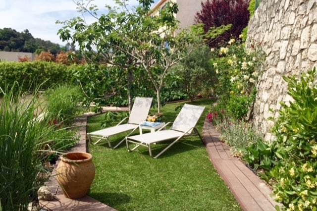 A serene garden space features two lounge chairs positioned on a grassy area, surrounded by lush greenery and colorful flowers. A stone wall provides a natural backdrop, while a tree and assorted plants create a calming atmosphere.
