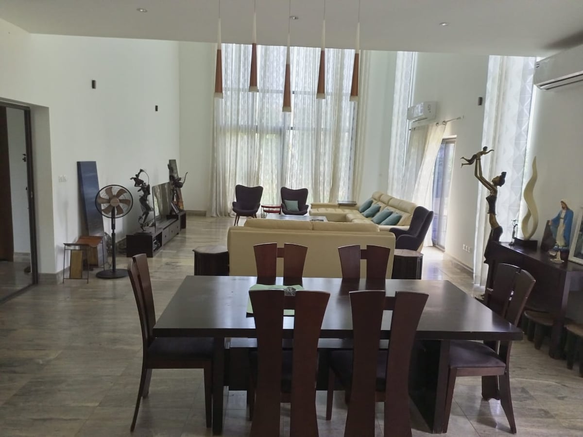 An open living space is highlighted by tall windows allowing natural light to fill the room. A dark dining table with multiple chairs is positioned in the foreground, while a comfortable seating area with couches and decorative sculptures can be seen in the background.