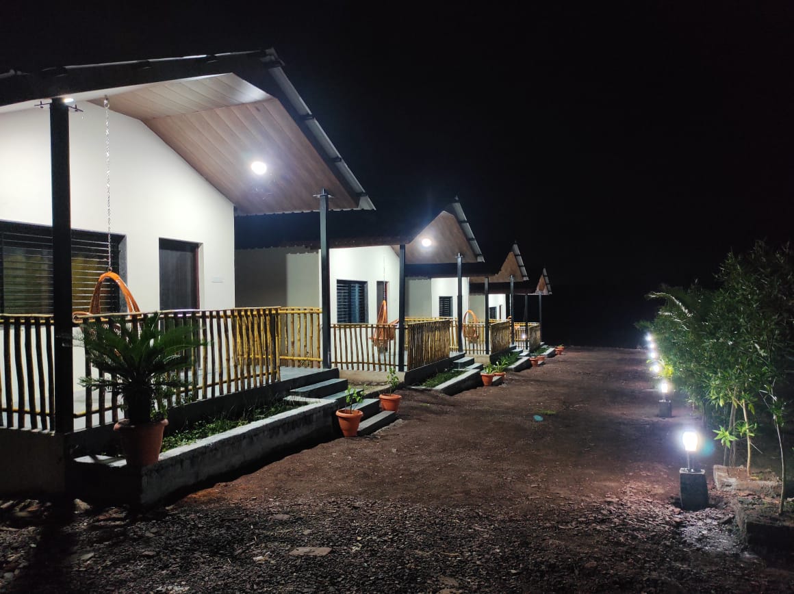 A row of charming cabins is illuminated by soft outdoor lighting at night, highlighting the welcoming facades and covered porches. Potted plants are placed along the pathway, creating a warm and inviting entrance to each unit.