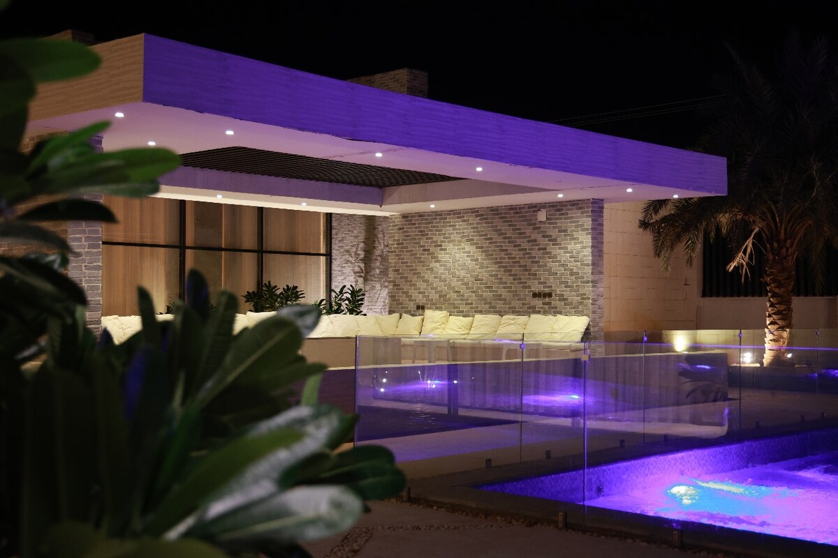 An outdoor lounge area is illuminated with soft purple lighting, showcasing comfortable seating arranged under a modern roof structure. Surrounding tropical plants add a natural touch, while the clear pool water reflects the evening ambiance.