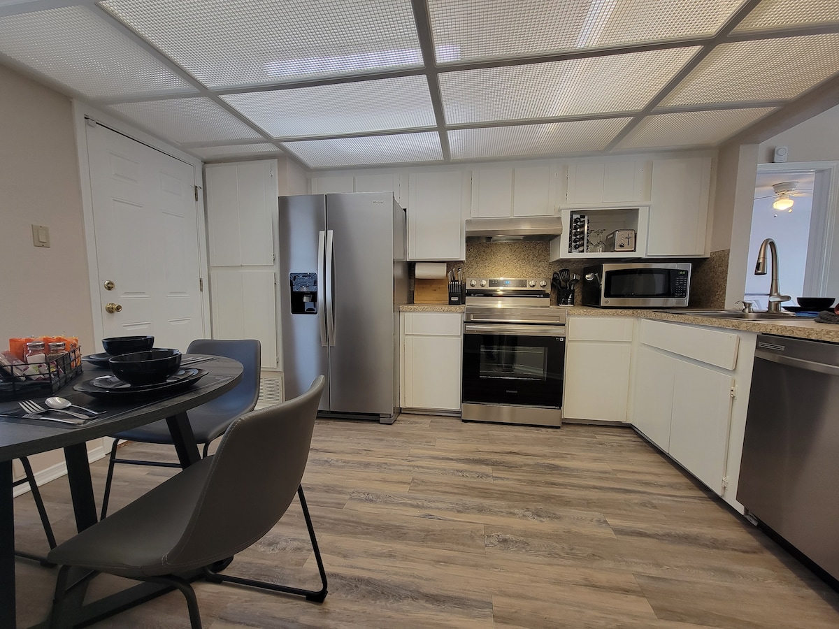 The kitchen features stainless steel appliances including a refrigerator, oven, and microwave. A spacious countertop provides ample preparation space. A dining table set for four is positioned nearby, complemented by simple grey chairs. The room is illuminated by natural light through overhead panels.