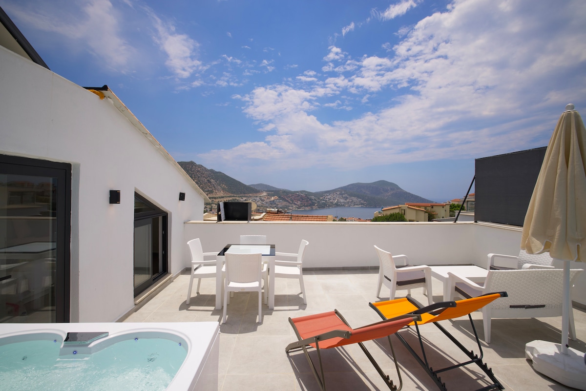 A spacious terrace features a hot tub, outdoor seating area, and sun loungers. Scenic views of the mountains and coastline are visible in the background, under a partly cloudy sky. The area is designed for relaxation and enjoyment.