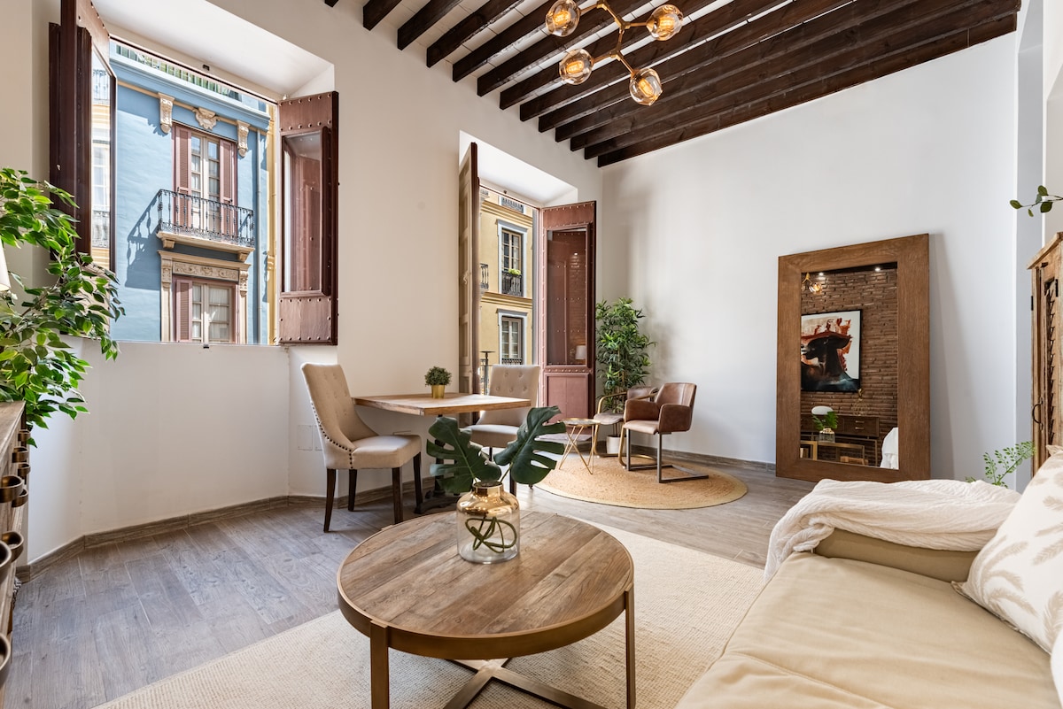 Natural light fills a cozy living area, featuring a comfortable sofa and a coffee table made of wood. Two large windows with wooden shutters open to a view of the vibrant neighborhood. A small dining table with two chairs is positioned beside a decorative wall mirror.