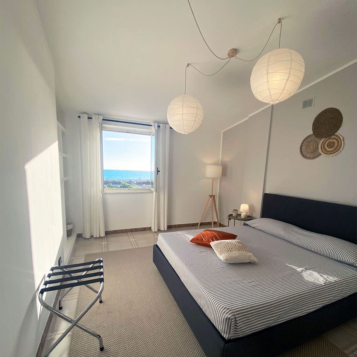 A well-lit bedroom features a spacious bed with striped bedding, complemented by a decorative pillow. Large windows offer a view of the sea, while a stylish lamp and a small folding chair complete the inviting space. Wall art adds a touch of character to the room.