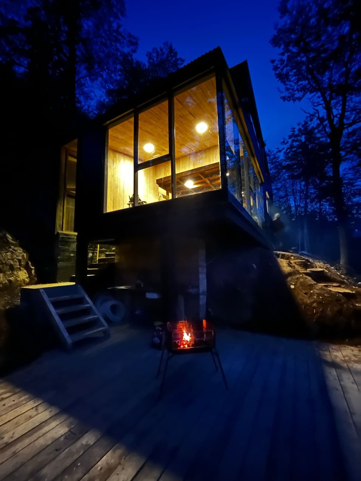An elevated cabin is illuminated by warm lights, showcasing a wooden interior and expansive glass walls. A fire pit, with glowing embers, is positioned on the wooden deck in front of the structure, surrounded by trees and rocky terrain, creating a serene outdoor atmosphere.