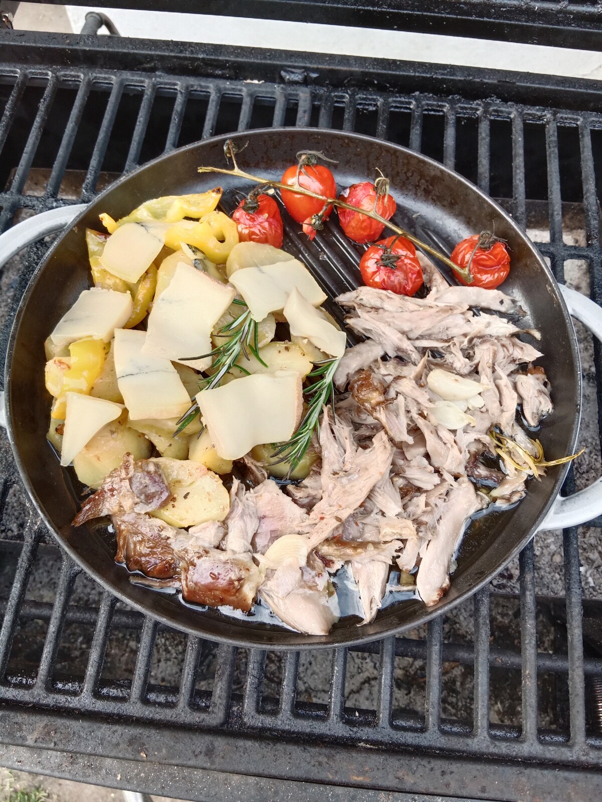 A round, black skillet is filled with various dishes, featuring pieces of shredded meat alongside golden potatoes and yellow peppers. Cherry tomatoes, roasted on the vine, add a pop of color, while fresh rosemary sprigs are visible, enhancing the visual appeal of the meal.
