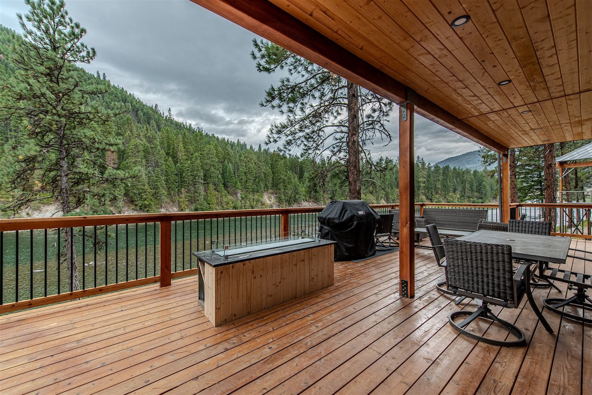 A spacious deck is shown with smooth wooden flooring, offering views of the lush green landscape and calm waters below. A sleek fire pit is positioned at the center, while seating arrangements and a grill are visible, providing an inviting space for gatherings.