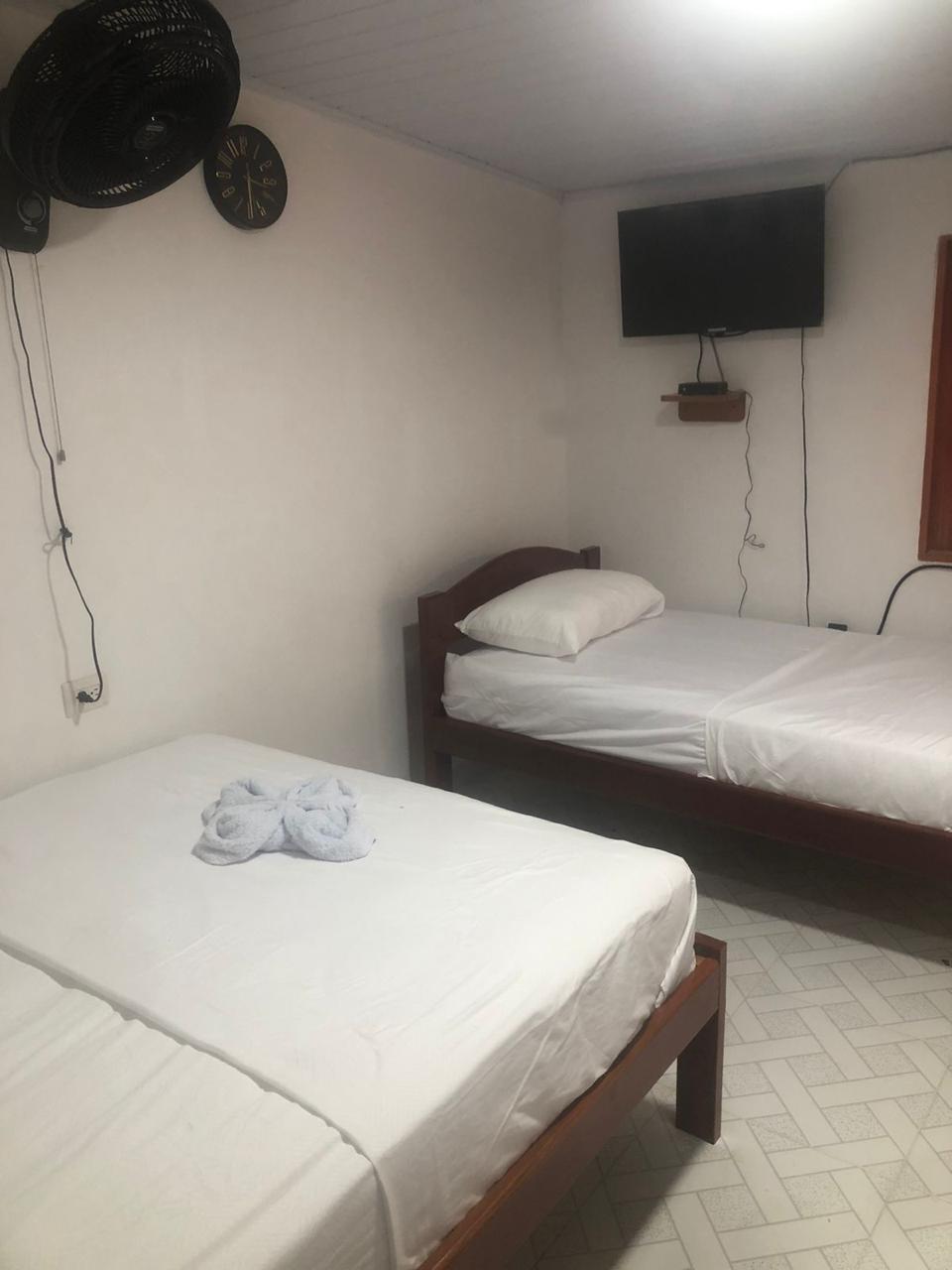 A simple sleeping area features two beds with white linens. A ceiling fan hangs above, and a flat-screen television is mounted on the wall. Natural light enters through a window with a wooden frame, providing a calm atmosphere.