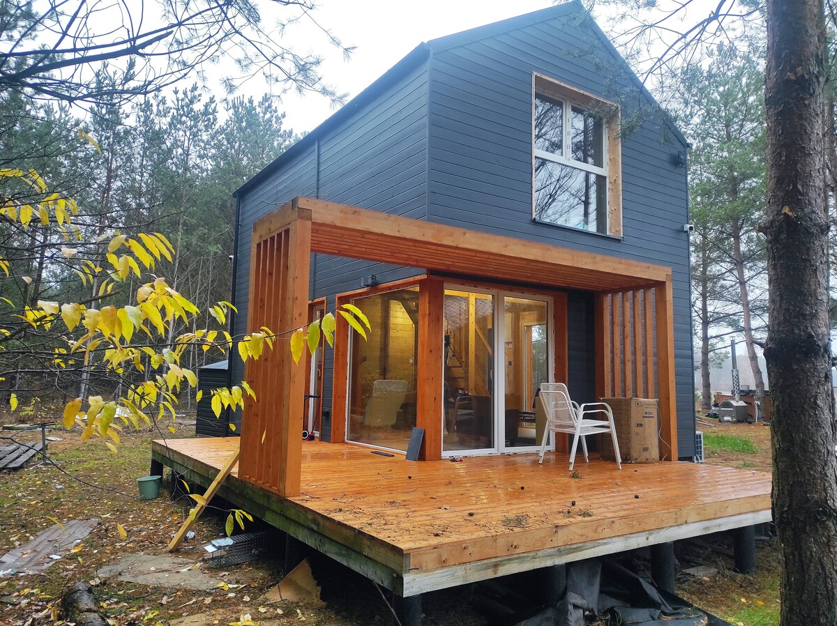 The exterior of a wooden two-story house is shown, featuring a large deck with wooden flooring. Two white chairs are positioned on the deck, inviting relaxation. Large glass doors provide views into the interior, surrounded by trees and nature.