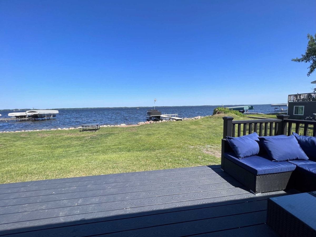 A grassy area leads down to Lake Osakis, with calm waters visible under a clear blue sky. A black outdoor seating arrangement with deep blue cushions is positioned on a deck, offering views of the lake and nearby docks.