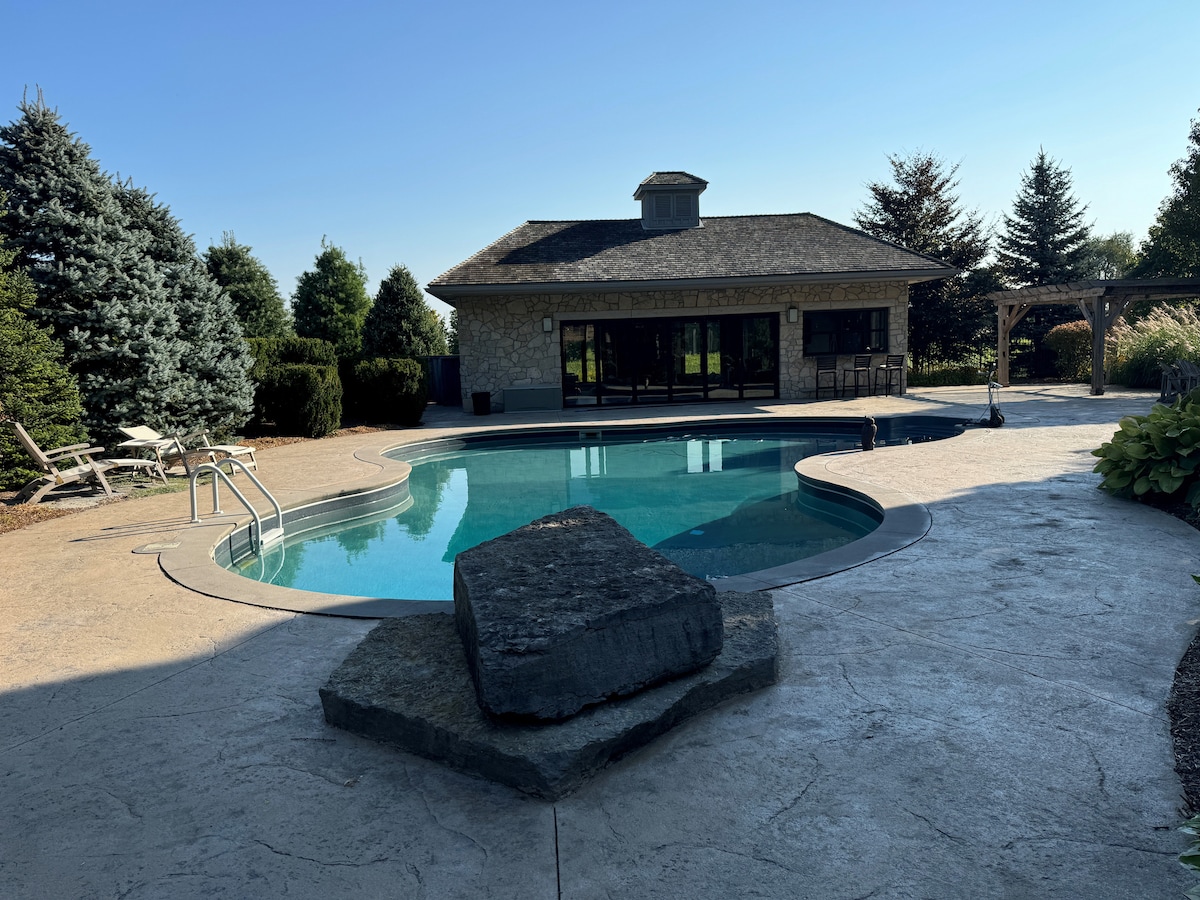 A private pool is framed by a spacious patio and surrounded by well-maintained greenery. The villa stands nearby, featuring large windows that overlook the serene outdoor area. A natural stone sits on the patio, adding to the tranquil ambiance.