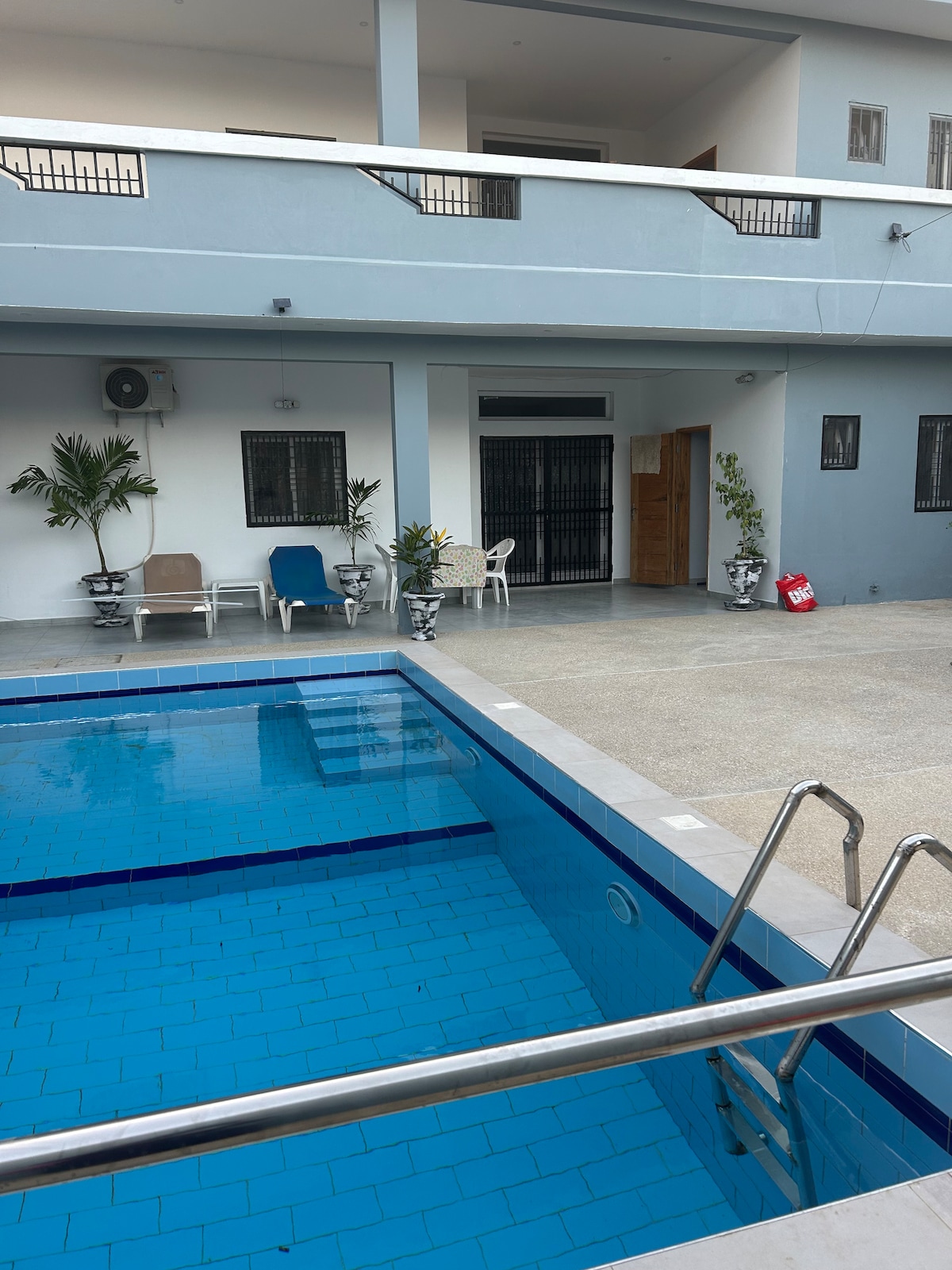 A clean swimming pool is framed by a concrete patio. Relaxing seating options, including lounge chairs, are positioned nearby. The two-story accommodation is visible in the background, with a balcony providing additional outdoor space. Potted plants add a touch of greenery to the scene.