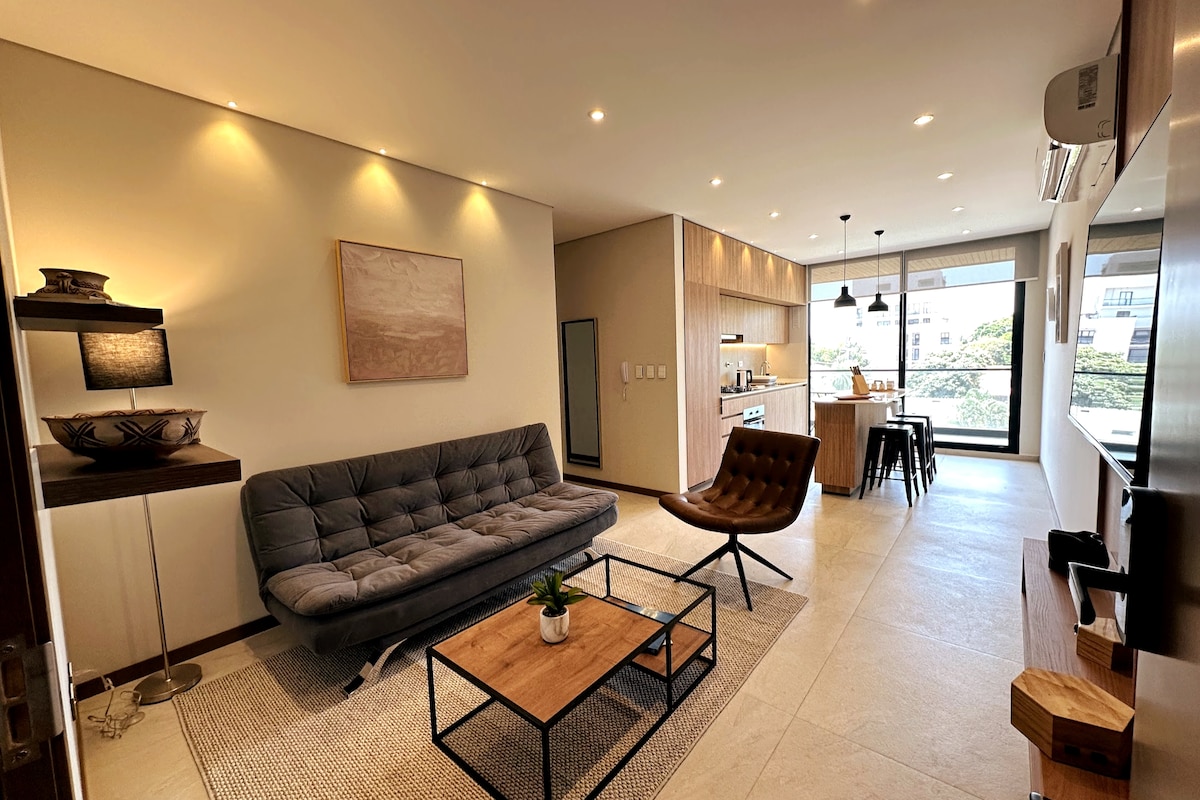 A modern living area is furnished with a gray sofa and a brown accent chair. A wooden coffee table sits on a textured rug in front of the sofa. Natural light floods in through large windows, illuminating the open layout that connects to an equipped kitchen.
