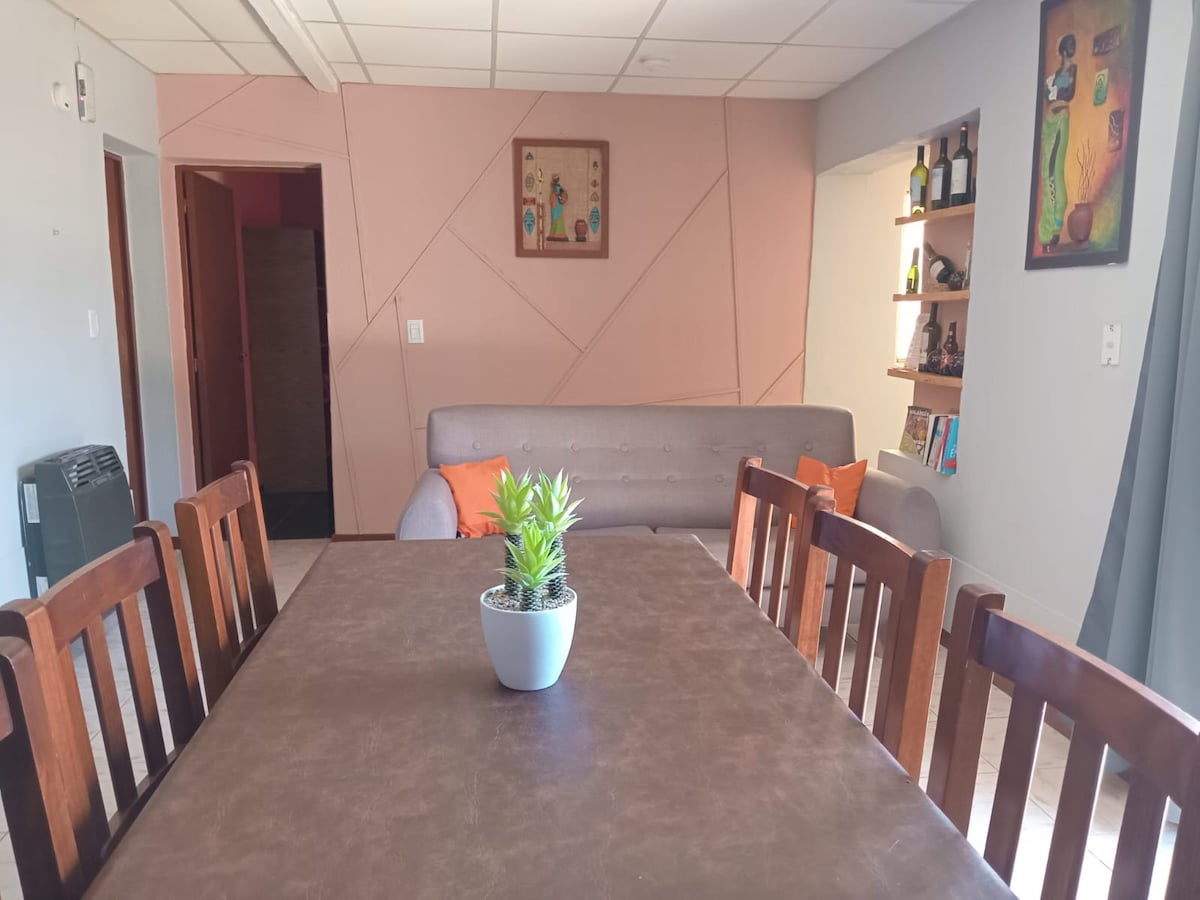 A dining area is featured with a large table surrounded by wooden chairs. A small potted plant sits in the center of the table. The room includes a gray sofa with orange cushions and decorative wall art, creating a comfortable and inviting space.