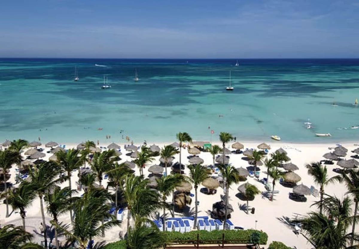 A panoramic view of a beach stretches across the foreground, featuring a sandy shoreline lined with thatched umbrellas and lounge chairs. The clear turquoise water extends into the distance, dotted with sailboats and gentle waves under a clear blue sky.