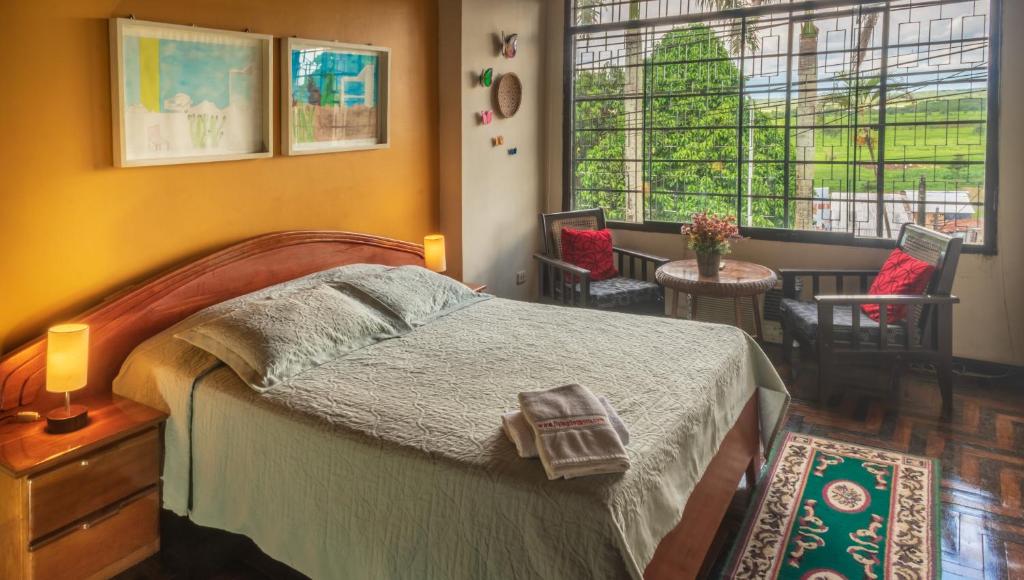A well-lit private bedroom features a spacious bed with light bedding. Two wooden chairs and a small table are positioned near a large window, offering views of greenery outside. Decorative wall art and soft lighting elements enhance the inviting space.