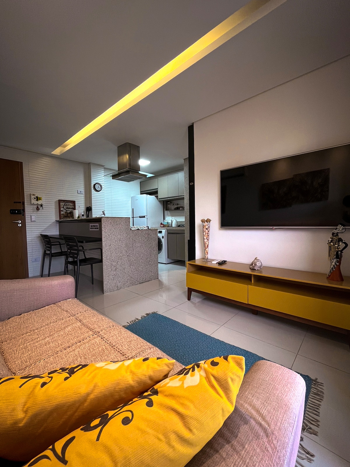 A modern living area is depicted with a cozy sofa adorned with decorative pillows. A large television is mounted on the wall opposite a stylish sideboard. The open-plan design reveals a sleek kitchen area featuring a countertop and essential appliances, bathed in soft overhead lighting.