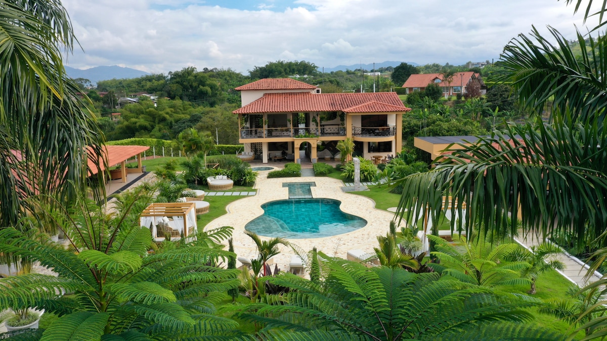 An expansive outdoor area features a large swimming pool surrounded by lush greenery and neatly arranged lounge chairs. The main building showcases a two-story structure with a red tile roof, complemented by shaded outdoor seating arrangements under wooden pergolas.