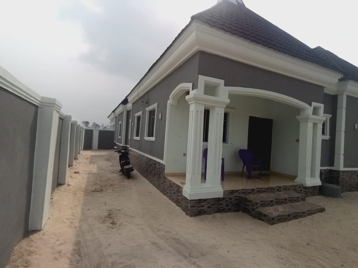 A modern house exterior is displayed, showcasing a structured porch with classic columns. The walls are painted in a subtle gray tone, complemented by a patterned stone base. An outdoor path leads to the entrance, and two purple chairs are positioned on the porch.