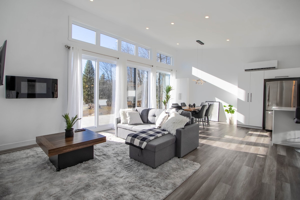A bright living area features large windows that allow natural light to fill the space. A sectional sofa is accented with a cozy throw blanket, while a wooden coffee table sits atop a plush area rug. A dining table for six is visible in the background.