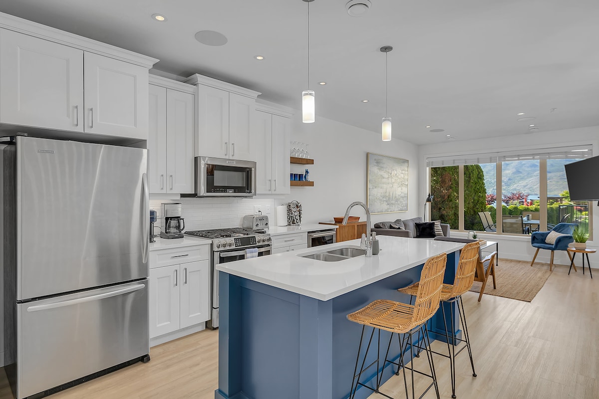 A modern kitchen space features white cabinetry, stainless steel appliances, and a large island with blue accents. Two stylish wicker bar stools are positioned at the island. Natural light floods the area through sliding glass doors, revealing a view of the outdoor space.