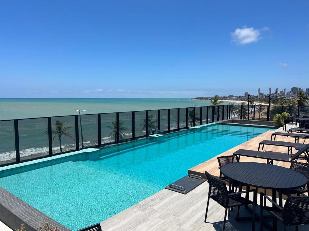 A rooftop pool area features clear turquoise waters, surrounded by a spacious deck with black outdoor furniture. The backdrop includes a stunning ocean view, with gentle waves and a sandy beach visible in the distance under a blue sky.