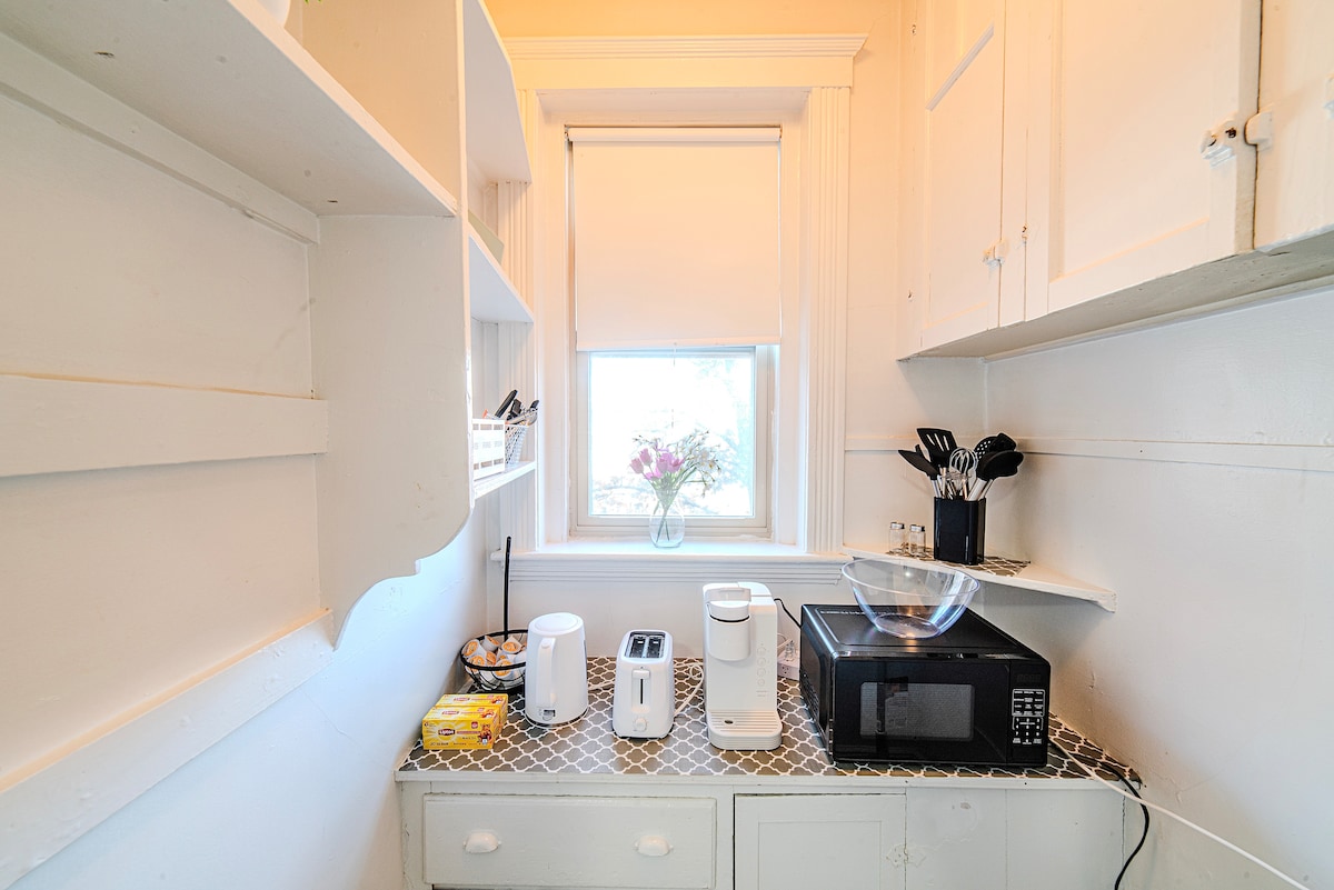 The kitchen features a functional layout with essential appliances, including a microwave, toaster, and coffee maker. A bowl is placed on the countertop, and natural light enters through the window, highlighting the neat organization of utensils and a floral arrangement.