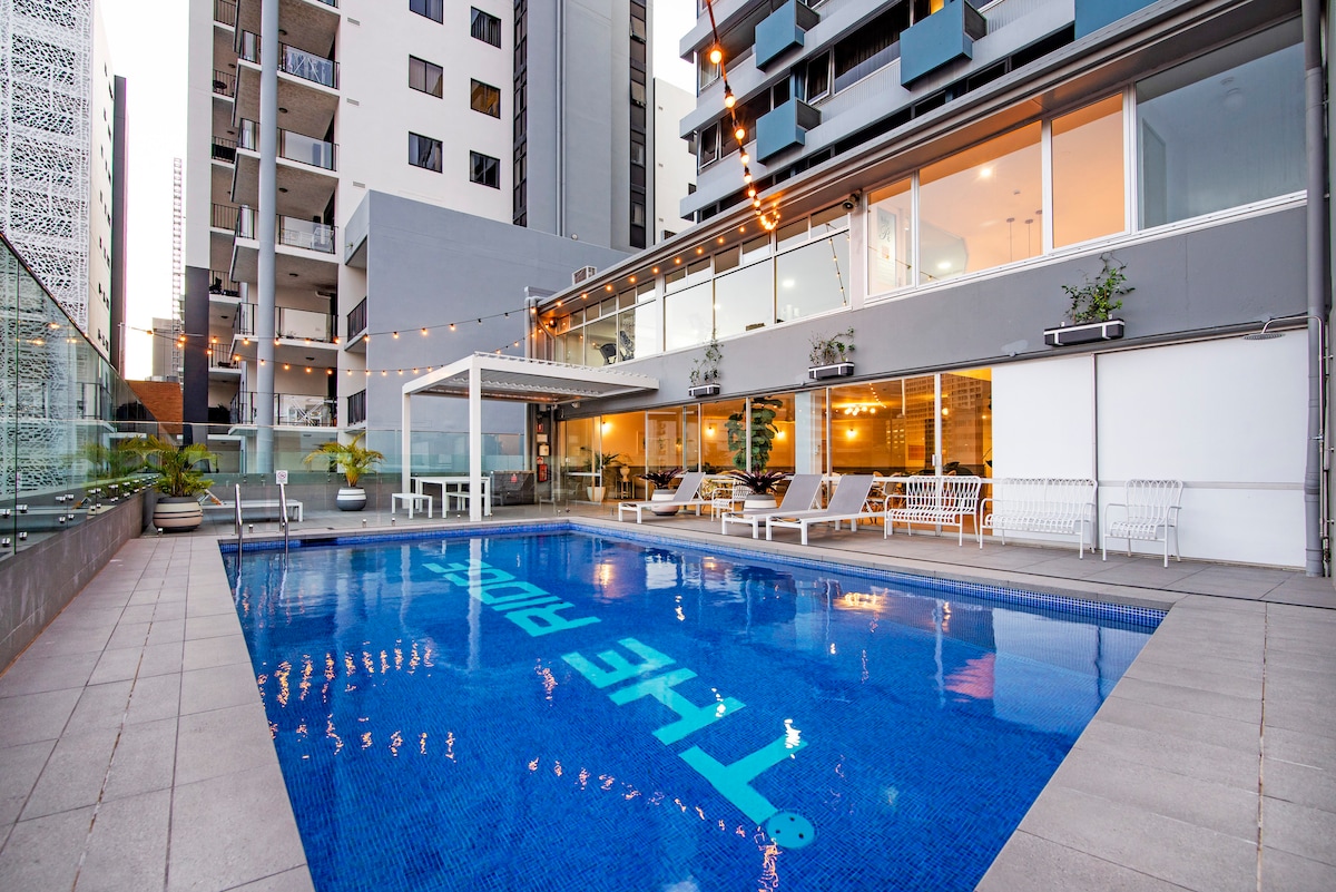 An inviting swimming pool is surrounded by a spacious area featuring stylish lounge chairs and modern outdoor furniture. Warm, ambient lighting adds a relaxing touch in the evening. The contemporary building structure with large windows is visible in the background, enhancing the open atmosphere.