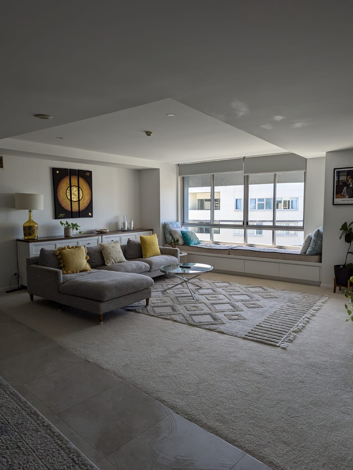 A stylish living area is showcased, featuring a comfortable gray sofa adorned with yellow cushions. A glass coffee table sits in front, and large floor-to-ceiling windows allow natural light to fill the space, complemented by a decorative rug on the light-colored floor.